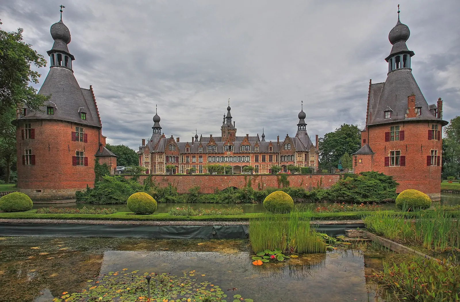 Het rode bakstenen Kasteel van Ooidonk met torens met uivormige spitsen, gezien over een vijver met waterplanten, tussen de twee torens van het poortgebouw.